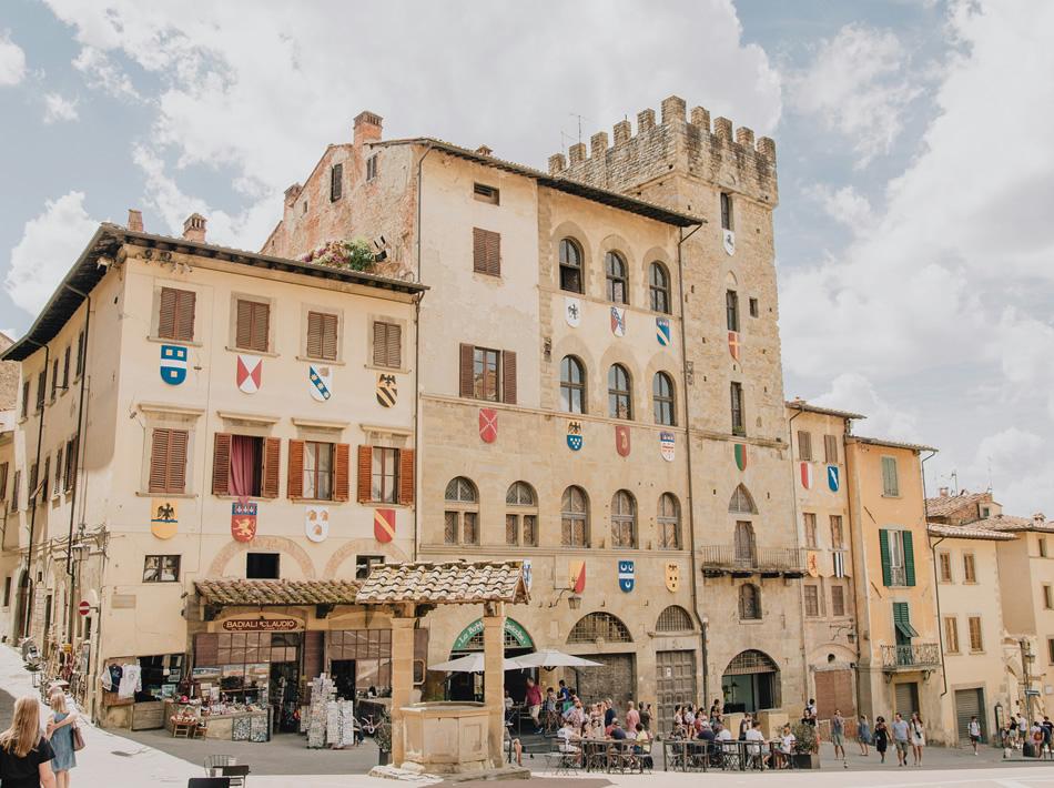 Piazza storica con edifici medievali e stemmi araldici.