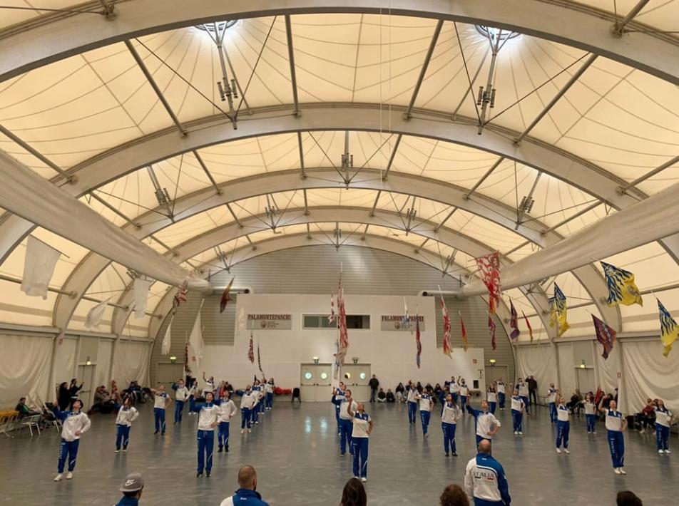 Esibizione di ginnastica ritmica in un palazzetto coperto con pubblico seduto ai lati.
