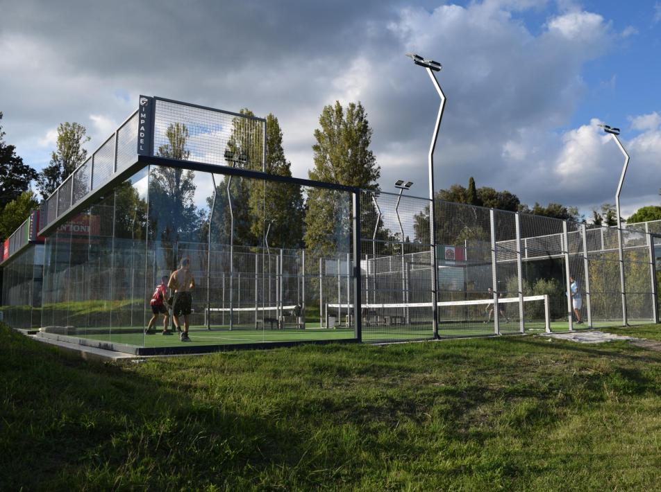 Giocatori di padel in campo all'aperto circondato da recinzioni, cielo nuvoloso.