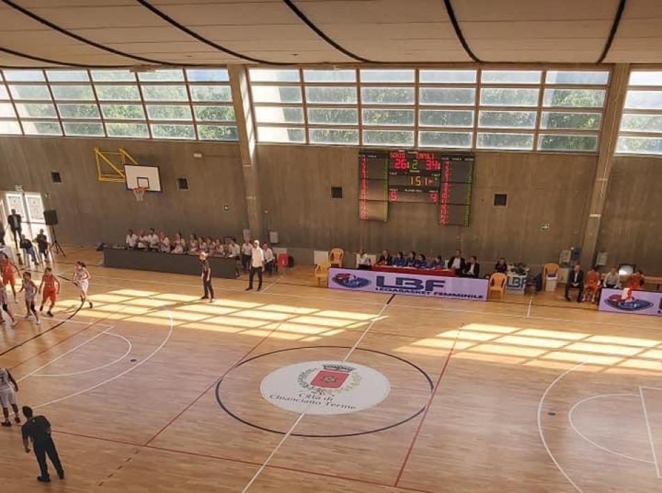 Partita di basket in un palazzetto con spettatori e tabellone segnapunti.