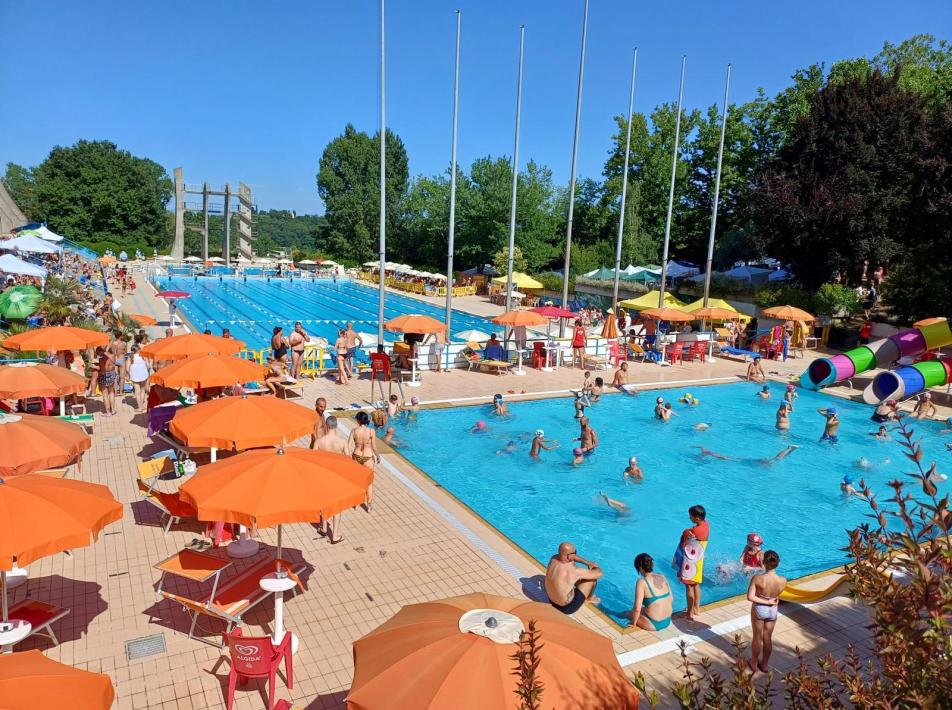 Piscina all'aperto affollata con ombrelloni arancioni e scivoli colorati.