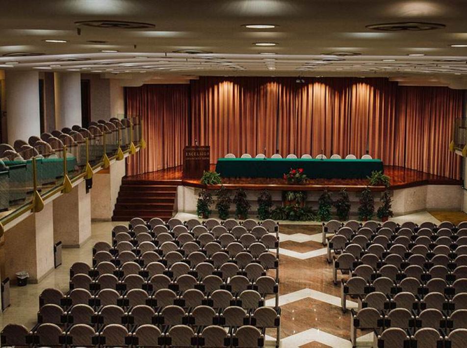 Sala conferenze con palco, sedie e tavolo per relatori.